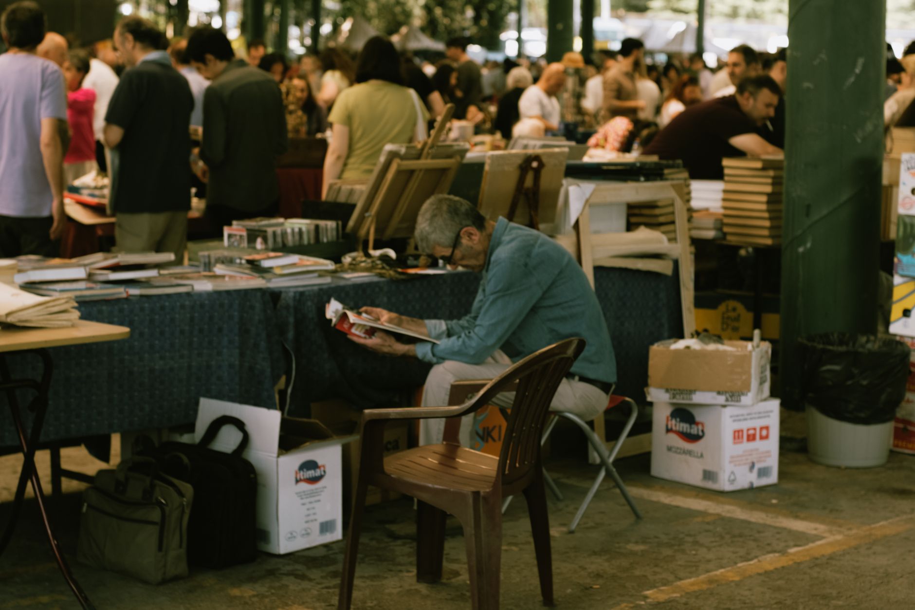 Older man reading a magazine at a bustling outdoor book market.