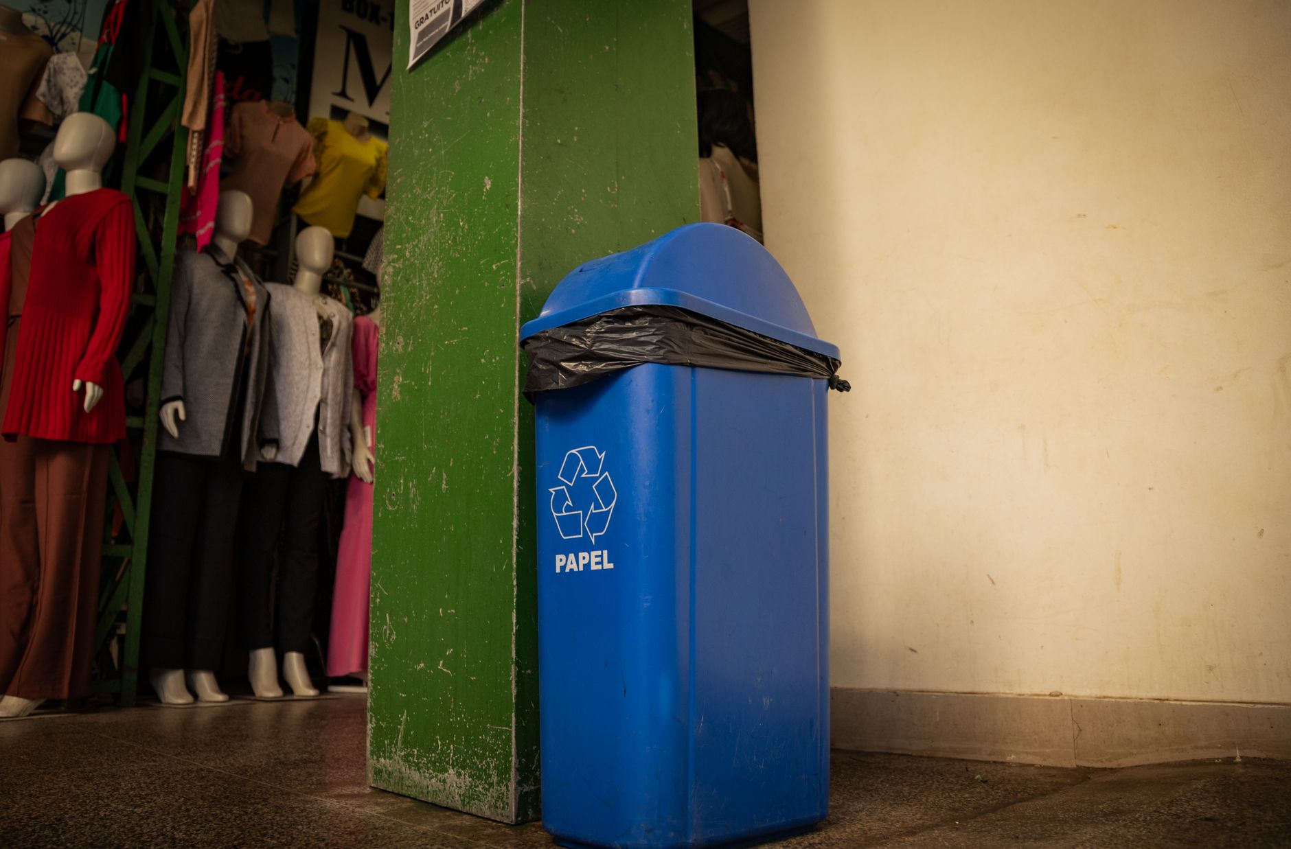 Blue recycling bin labeled 'papel' inside a boutique with mannequins and clothing.