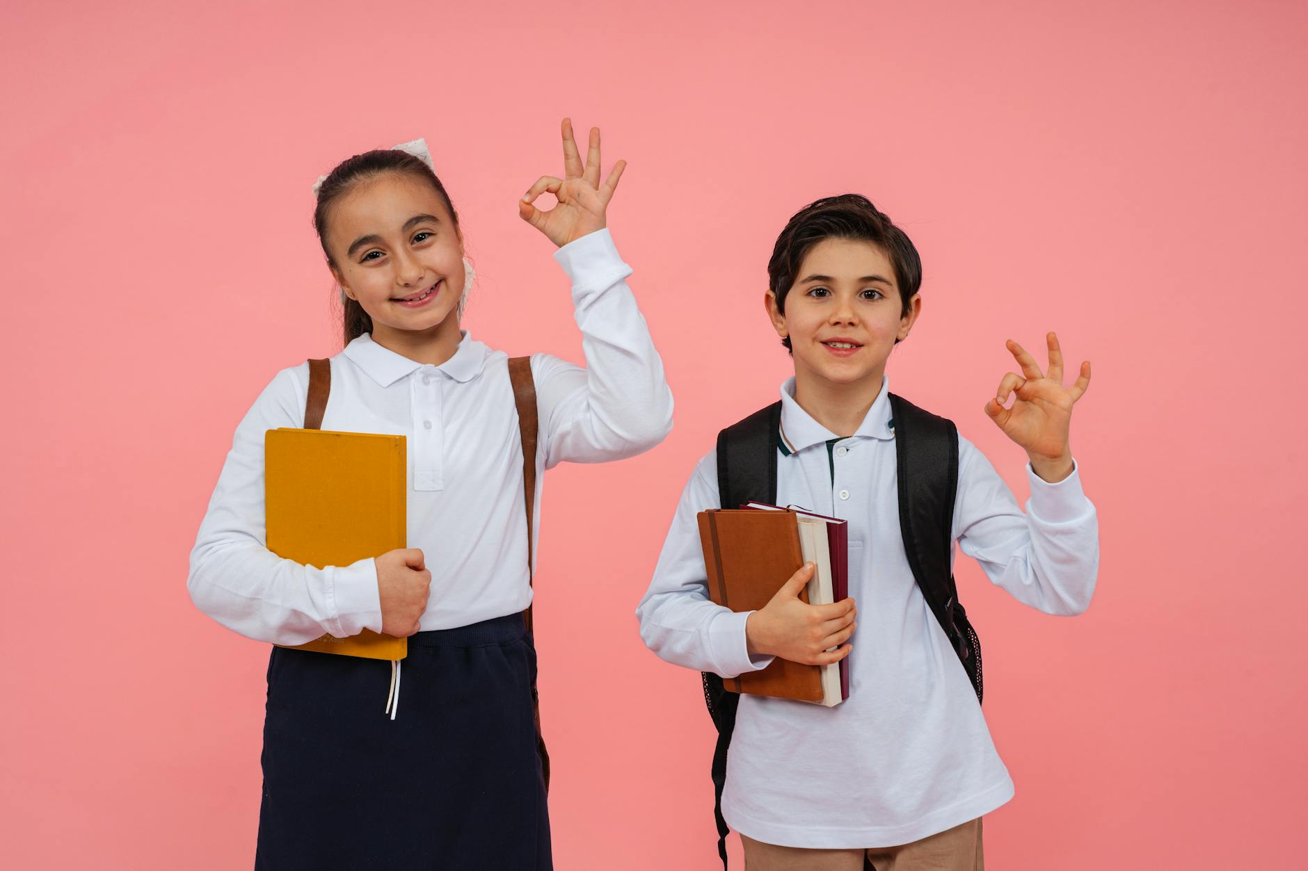 Two cheerful schoolchildren giving OK sign while holding books against a pink background, Yalova.