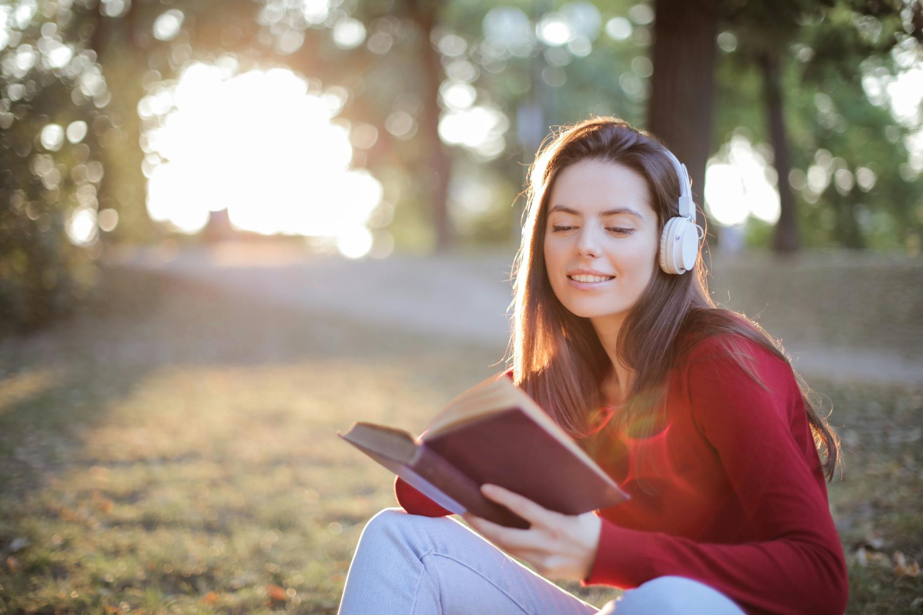 A woman wearing headphones reads a book outdoors, enjoying a sunny day in the park.
