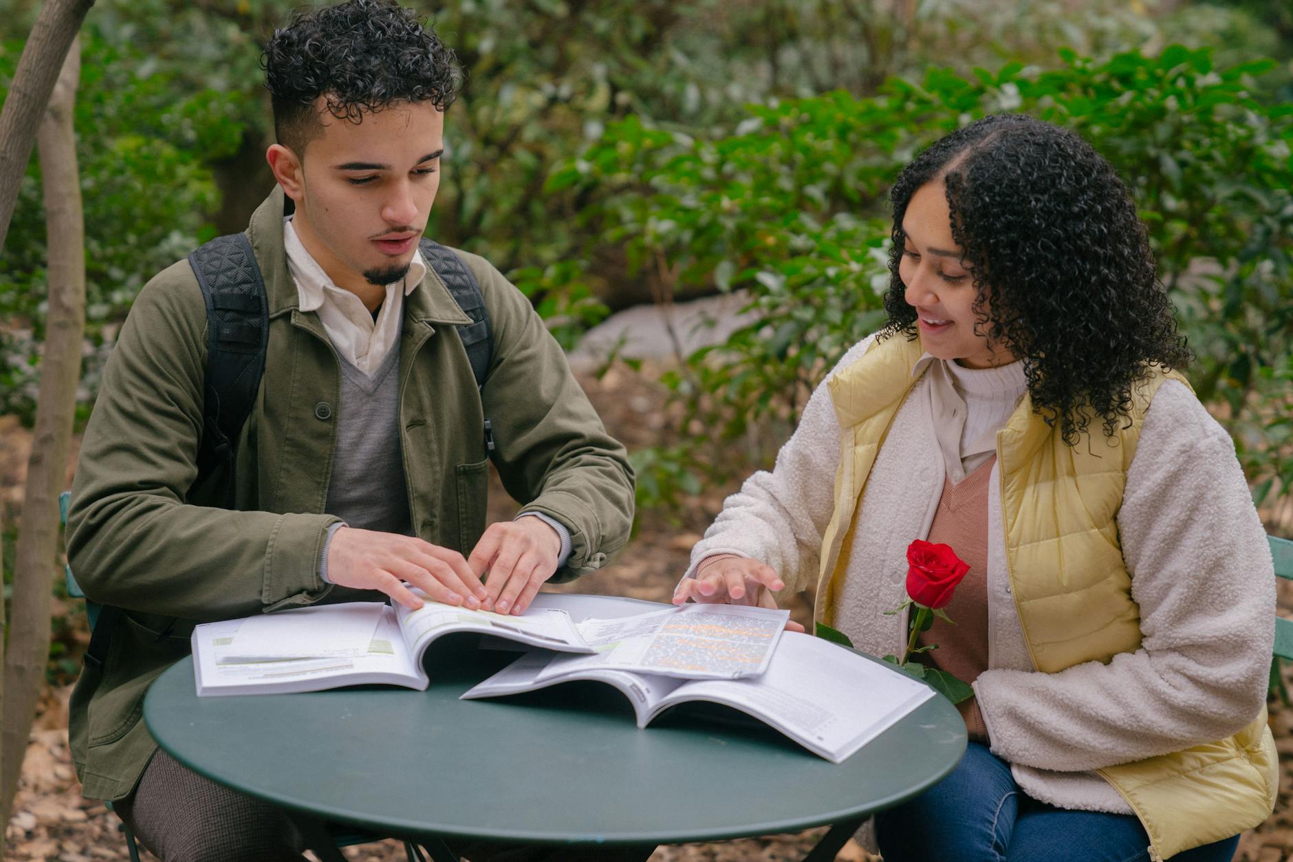 Young Hispanic couple in warm clothes with red rose reading textbooks while studying at table on chair near green bushes and trees in park in daytime