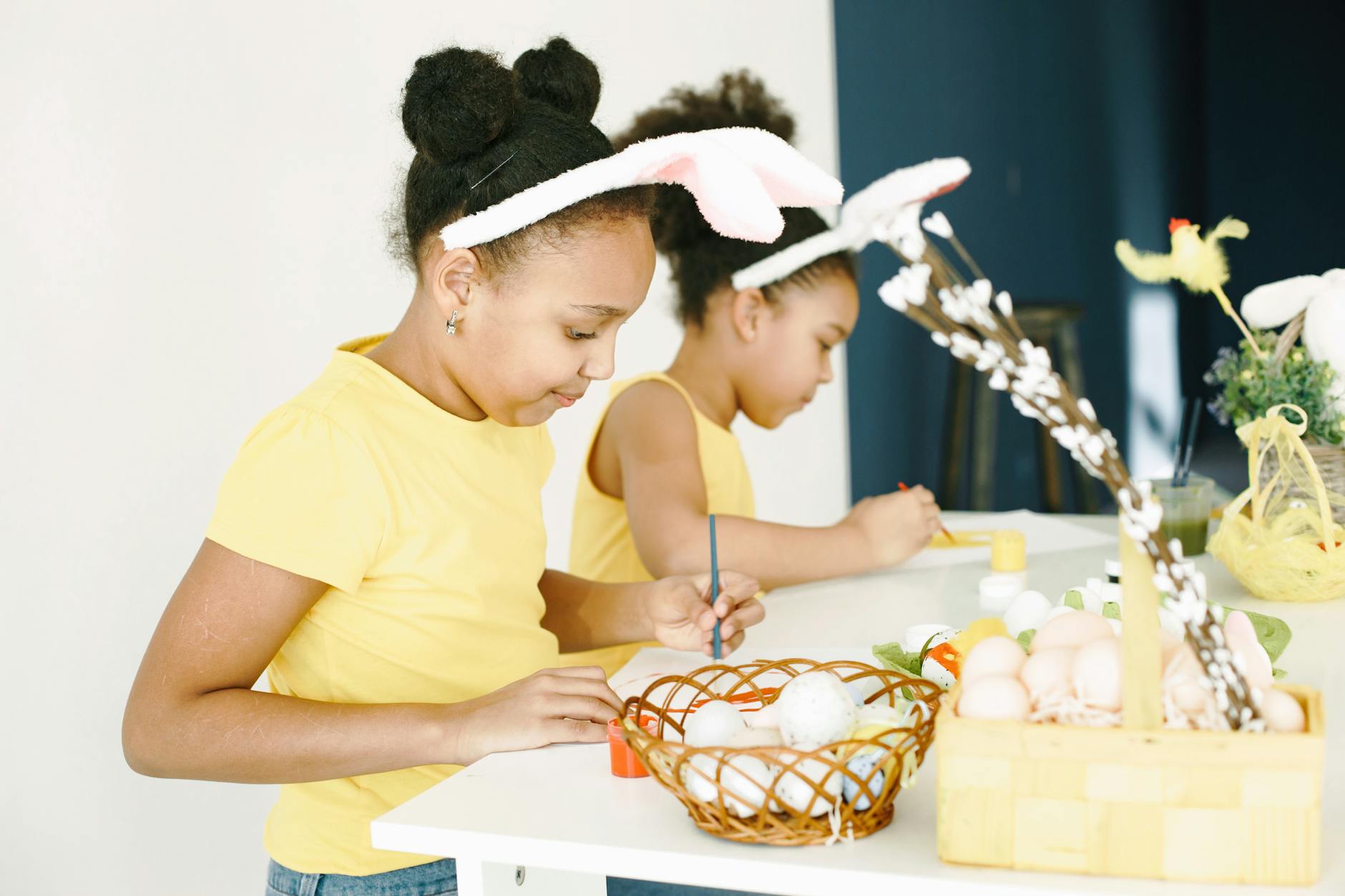 Girls painting Easter eggs indoors, wearing bunny ears and yellow outfits, having fun.