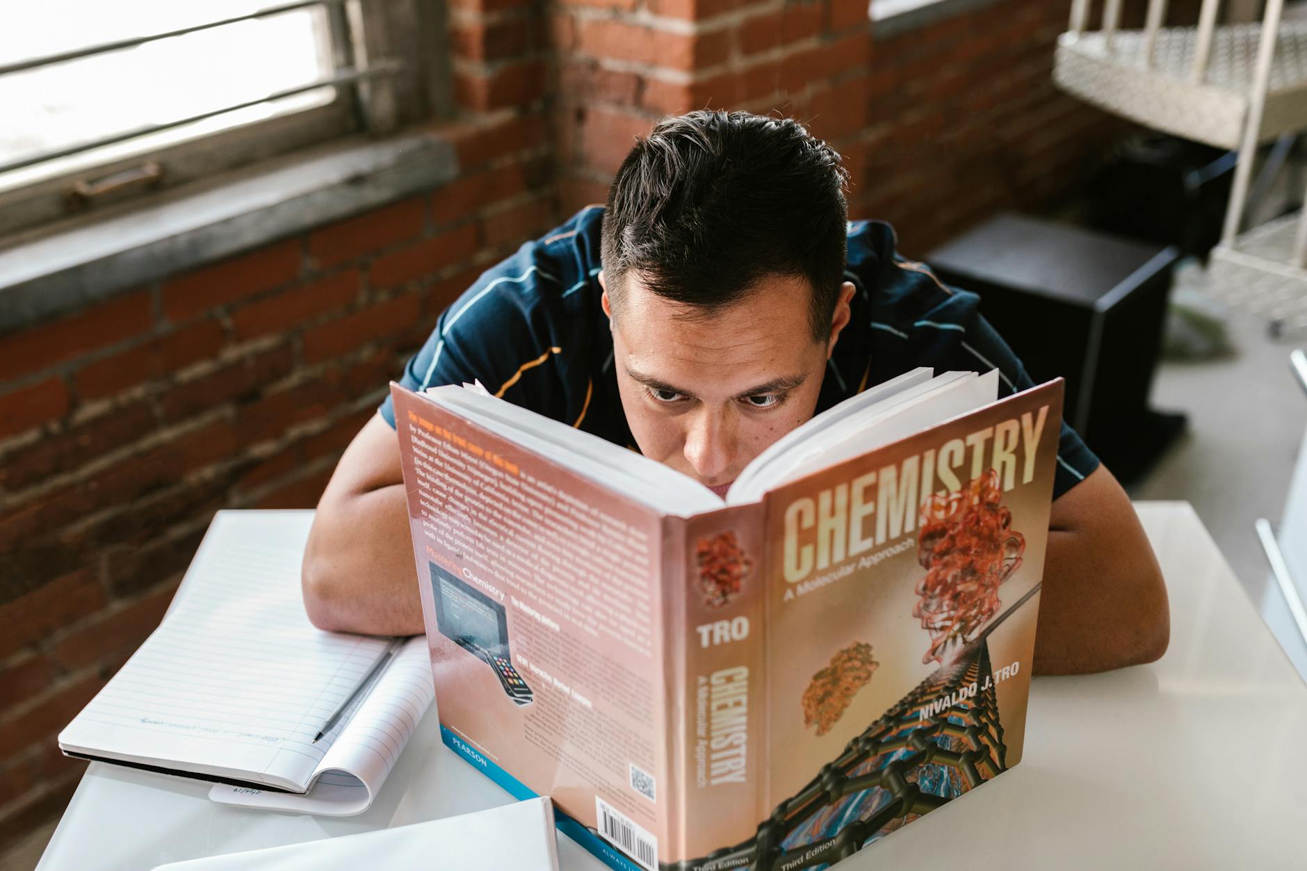 Adult male studying a chemistry textbook at a desk in a cozy, brick-walled study area.