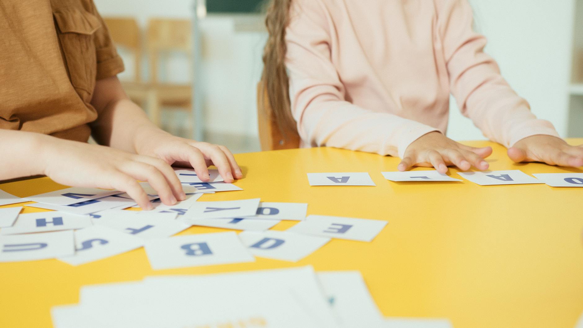 Children arranging alphabet cards on a yellow table in a classroom setting.