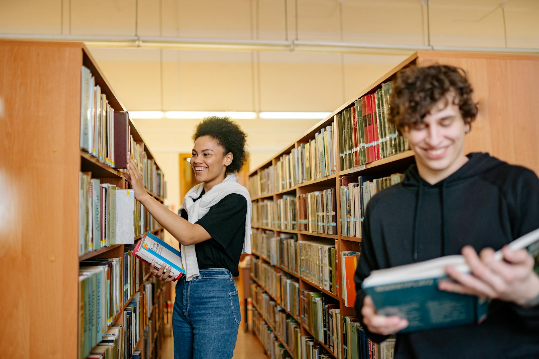 Two students studying in a university library, choosing books from shelves.