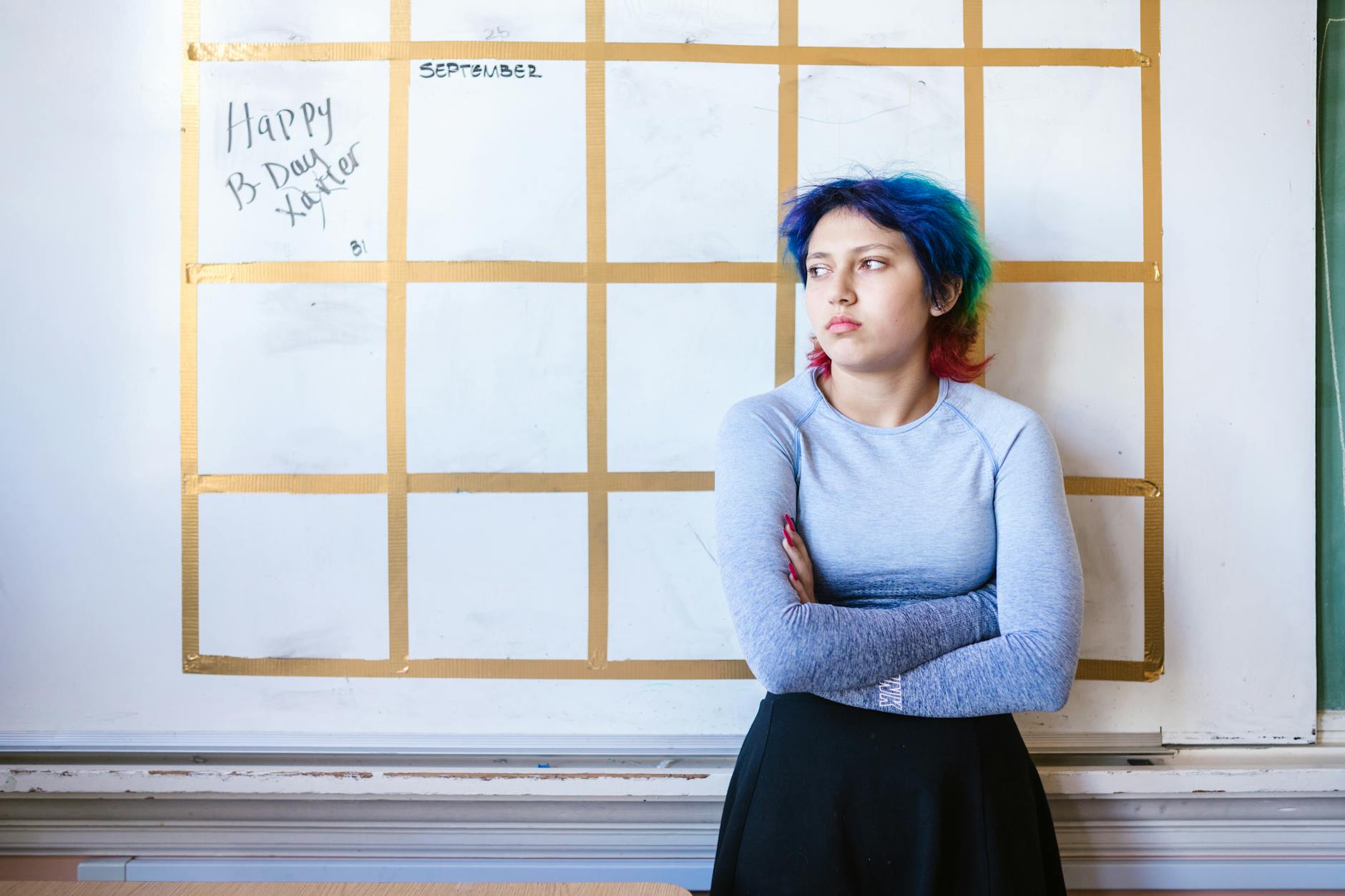 Teen with colorful hair stands pensively in front of a school whiteboard.