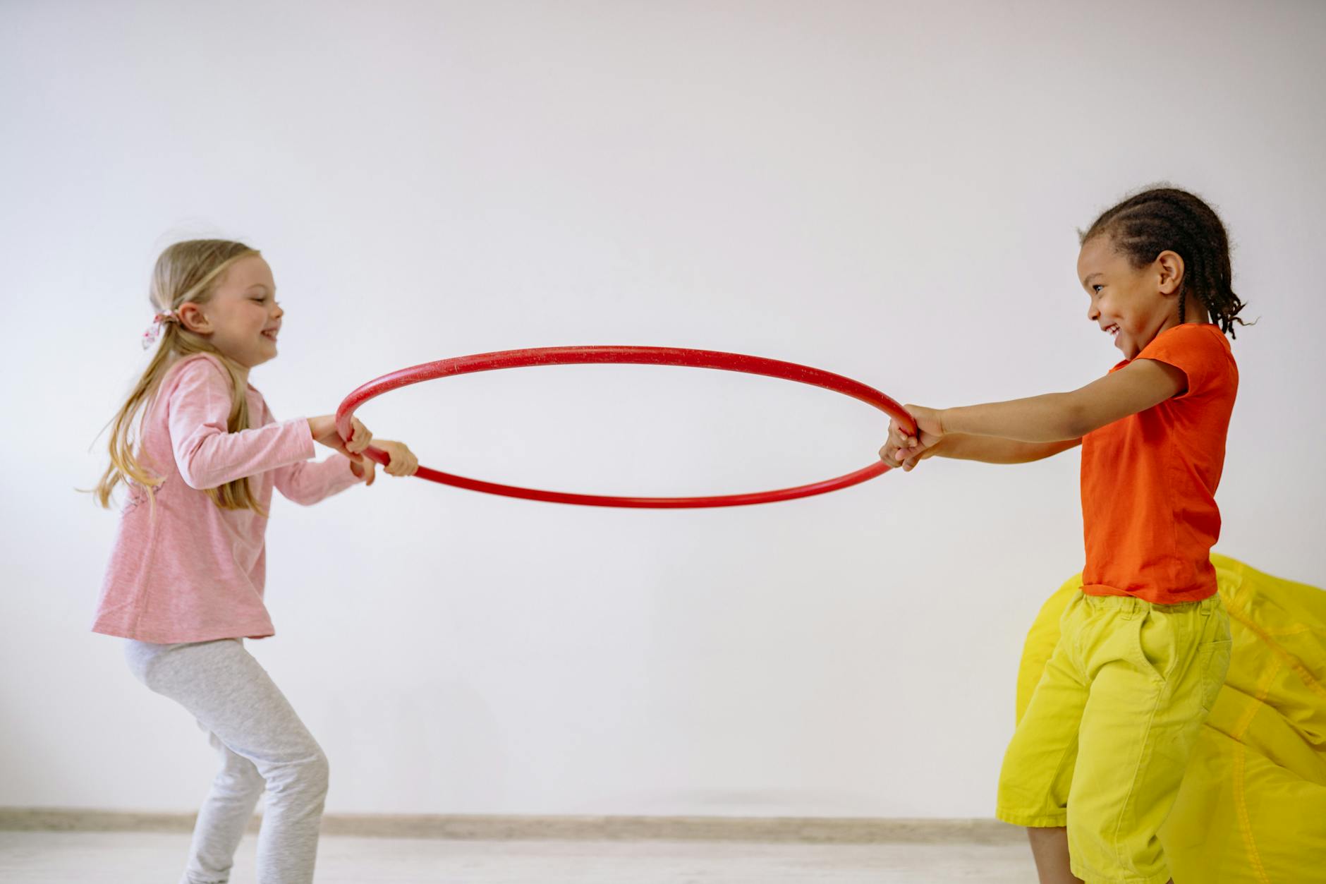 Two children joyfully playing with a hula hoop indoors, sharing laughter and fun.
